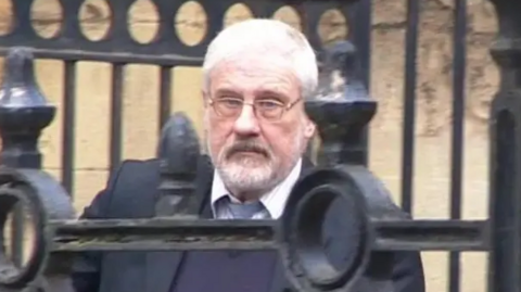 Stephen Cox, wearing a buttoned-up shirt and tie, but with the top button undone, and a purple jumper and dark jacket, walking out of Reading Crown Court, with the bars of railings in front of him as he walks down a ramp outside the building.