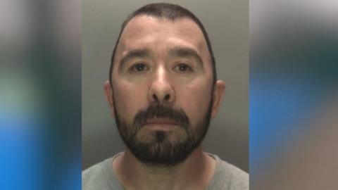 A mugshot of a man in a grey t-shirt. He has a dark beard and short dark hair.