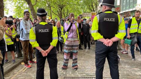 Police with their backs to the camera holding a line in front of a large number of counter-protesters. One woman is shouting into a megaphone in front of them. 