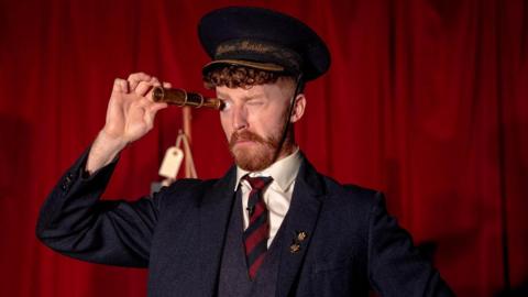 An actor dressed in a three-piece suit looks through a monocular. Behind him is a red curtain. 