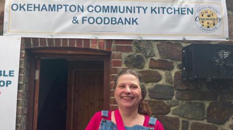 The founder of Okehampton Community Kitchen and Foodbank, Rebecca Green, standing outside the doorway of a red brick building. She is wearing mid-blue denim dungarees with pink hearts embroidered all over them, and a fuchsia pink knitted three-quarter sleeve top underneath.