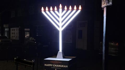 A street scene at night with a large menorah , which is the Jewish candelabra, with 9 bulbs on top. The stem and V-shaped prongs are lit up and glowing white. It stands on a black box with the words Chabad Bricket Wood wishes you a Happy Chanukah. Behind it are houses with cars on their driveways. 
