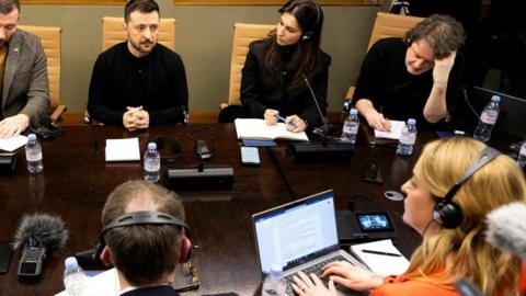 Zelensky sits at a table surrounded by journalists wearing headphones and typing on laptops