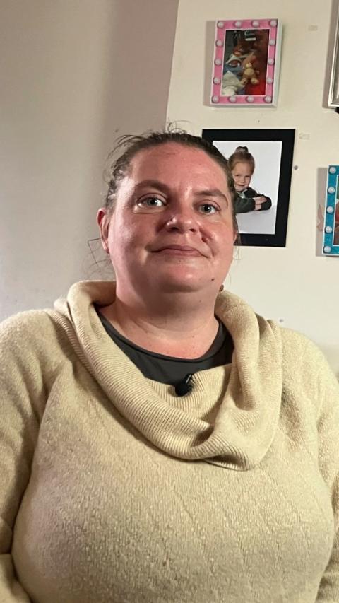 A woman smiles gently while posing for a photo. She is in her home and there are photos on the wall behind her. She is wearing a beige jumper with a rolled neck.