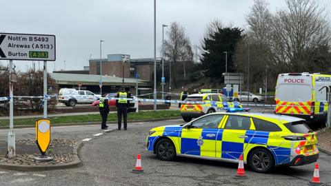 Police cars and a police van and cordon outside the station. At the front of the picture, a police car with blue and yellow markings is positioned and there are traffic cones on the ground. Four police officers are also in the picture. 