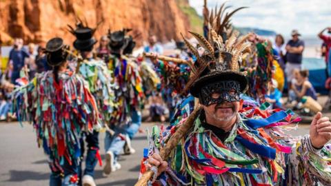 Morris dancers wearing multiple coloured rags and black top hats with feathers perform along Sidmouth seafront during the town's Folk Festival. The dancer nearest to the camera is wearing a black mask over his eyes. A crowd is watching the dancers.