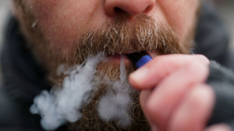 Close up of a man using a disposable vape. The man has a dark moustache and beard. The image shows a purple vape in his mouth. He is blowing smoke out of the corner of his mouth. He is holding the vape with one hand and is wearing fingerless gloves.