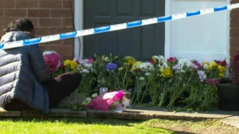 A woman wearing a blue coat and putting flowers outside the house. There is blue and white police tape across the scene