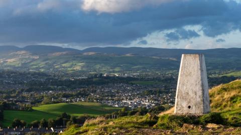 Trig pillar painted white can be seen sitting on a hilltop overlooking a town