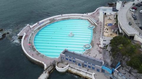 Drone shot of Tinside Lido in Plymouth. The base of the pool is light and dark blue stripes. An area of water called Plymouth Sound is to the left of the lido. There is a road above the lido with some cars parked on it