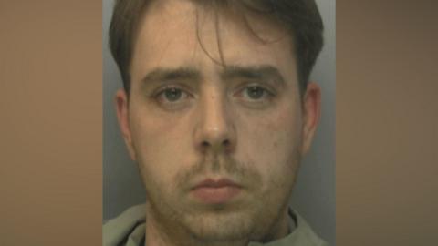 A mugshot of a man with medium-length brown hair. He has stubble and a grey jumper on.
