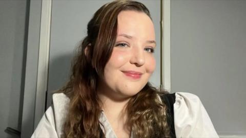 A woman with long brown hair wearing a white blouse smiles at the camera 