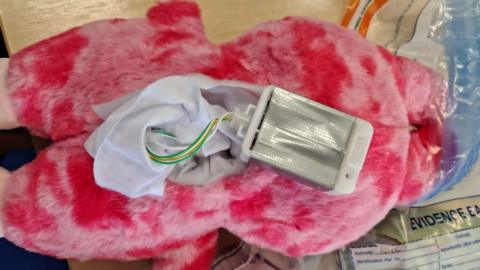 The back of a light and dark pink coloured furry toy with the battery pack exposed, showing green, white and yellow wires. It lies on a table top next to a clear plastic bag marked with the word 'Evidence'.