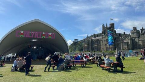 A covered stage sits on a field with a pink sign reading "Canlynliadau" meaning results in English. Margam Castle can be seen in the background on the right hand side. People sit on picnic benches in the sunshine outside the stage. 