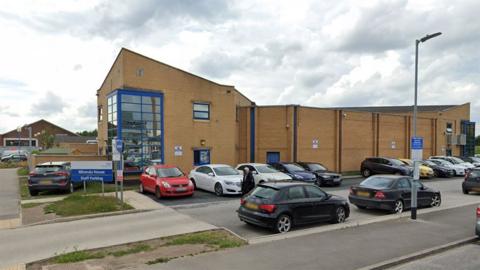 Google street view of Miranda House a two-storey building made from yellow brick with a large blue window at the corner and cars parked in front