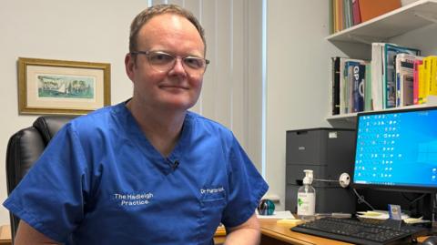 A GP in his fifties wearing glasses in his consulting room. 