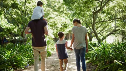 A family of four walks through a park with their backs turned to the viewer. The woman on the right is holding hands with a boy while the man on the left has a young girl on his shoulders.