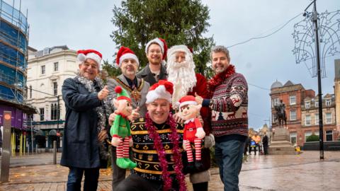 Six men stand in front of a Christmas tree in Wolverhampton town centre and smile at the camera. Five of the men wear Santa hats while one man is dressed in a complete Santa suit. The man kneeling at the front of the group holds up two toy elves.
