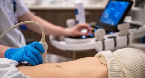 A medical professional performs an ultrasound examination on a patient, with a focus on the hand holding the transducer and the ultrasound machine screen in the background. The scan is being done on a woman's stomach, and the hand holding the equipment is covered in a blue glove.