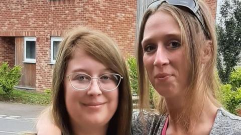 Two women standing outside a brick building. The woman on the right has her arm around the other woman. 