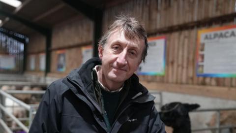 Dr Sean Taylor stands in a barn, stood against a gate wearing a dark jacket. Behind him in a pen can be seen a black alpaca.