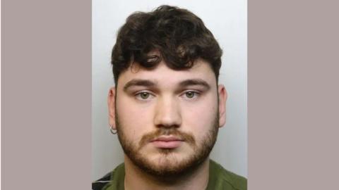 George Rogers looking into the camera for a police custody image with his left ear pierced, short dark brown curly hair and a beard