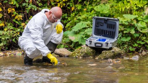 General image of a scientist in a white waterproof suit, with black boots and yellow gloves, testing the river water for pollution