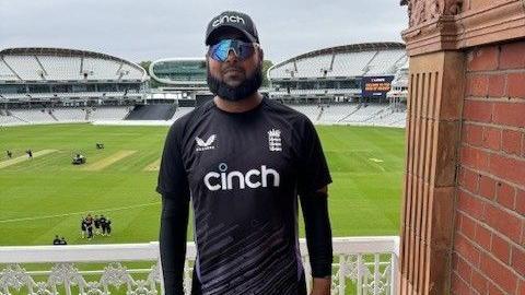 Moshfique Ahmed, a man with a black beard and wearing a black t-shirt and baseball camp and a pair of sunglasses, stands on a balcony with a cricket stadium behind him.