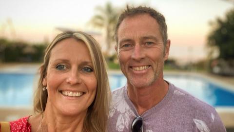 A man and woman smile on holiday. The woman has shoulder length blonde hair and is wearing a red top, the man has short brown hair, stubble and a lilac t-shirt. There is a pool behind them.