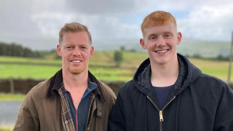 Two men stand next to each outdoors with green fields behind them. They are wearing coats and smiling.