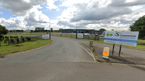 A road into Snetterton Business Park, showing entrance signage and road bollards, with parked cars and buildings in the distance. The sky is cloudy.