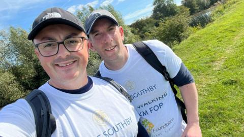 Dave Southby and his colleague Neil Sawyer posing for a selfie beside a river in a grassy field. They are both wearing caps and white t-shirts which say 'Dave Southby Financial Planning. We're walking 100km for the Kelly Foundation.'