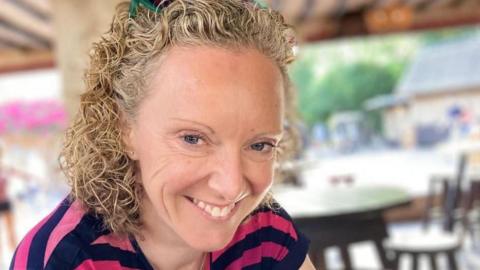 A woman with curly blonde hair smiles at the camera. She is wearing a stripy blue and pink T shirt.