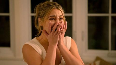 A still from I Know What You Did Last Summer shows actress Madelyn Cline with her hands clasped to her face, mid-scream. She's inside a house at night with large bay windows behind her.