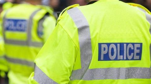 Three police officers who are standing outside and are wearing bright fluorescent yellow jackets which has grey stripes on the back and it says POLICE on the back. They all have their backs turned to the camera. 