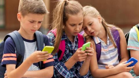 Three children, each holding mobile phones, sitting beside each other on a wall. To the left is a boy with short brown hair, wearing a grey t-shirt and holding a yellow-backed phone. Two girls with shoulder length brown hair holding green and blue backed phones are beside him.