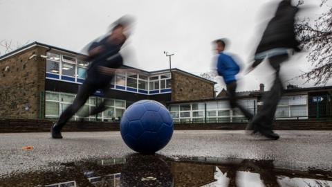 A photograph showing blurred images of school pupils playing football in a school playground. A school building can be seen in the background.