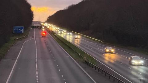 A motorway with vehicles travelling towards the camera on the right. There is no traffic on the left carriageway. The road is lined with trees and the sun is going down in the background