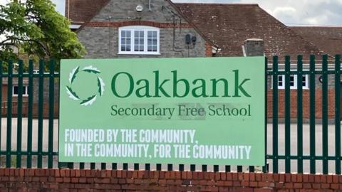 A green sign attached to metal railings displays Oakbank school's name. The green sign reads 'Oakbank Secondary Free School' with the slogan underneath saying: Founded By the Community, in the community, for the community'.  