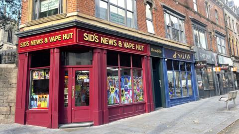 A street corner in Leeds city centre. The shop in foreground is painted dark red and has a sign which has "Sid's News & Vape Hut" written in cream. Other shops, including a salon (painted blue) can be seen to the right. 