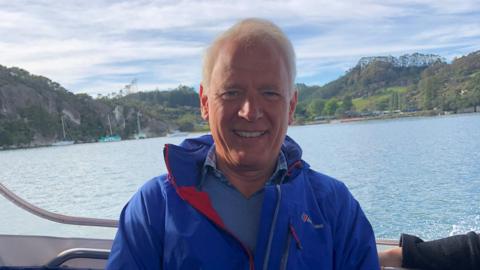 A man in a blue jacket is smiling while sat on a boat. Water and green hills can be seen in the background.