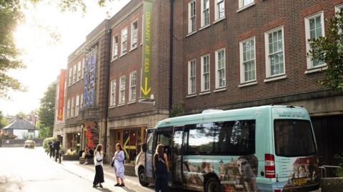 A blue shuttle bus outside a large building. There are passengers standing around the bus.