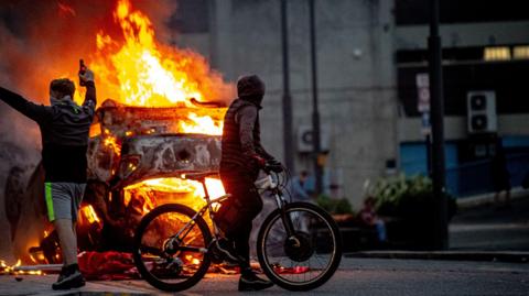 A burning car in the middle of the street. Large orange flames engulf the car which has been completely destroyed. A man wearing a hood and riding a bicycle has stopped to watch the flames. A man standing to his left has his arms in the air. He is holding a bottle and has a the lower half of his face covered.