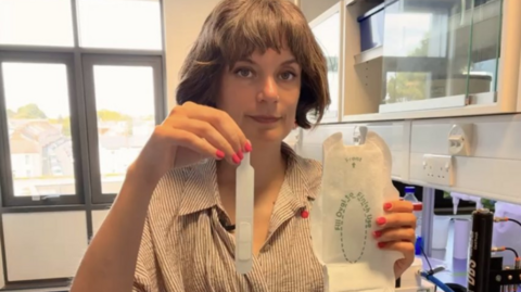 Professor Gemma Sharp, an epidemiologist at the University of Exeter is holding up a specialist sanitary pad and pipette and looking at the camera in a lab. She has short brown hair in a bob and is wearing a brown and white pinstripe blouse.