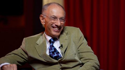 Mae Syr Michael Mortiz yn eistedd mewn cadair llwyd mewn ystafell â llenni coch yn y cefndir. Mae’n gwisgo siwt lliw olewydd gyda chrys gwyn a thei â phatrwm glas a du. Mae ei gorff wedi’i droi ychydig i’r ochr, ac mae un llaw yn gorffwys ar fraich y gadair tra mae’r llall yn llonydd ar ei goes.