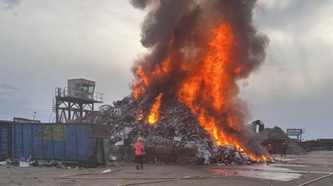 A firefighter dealing with a blaze at a recycling centre. A large plume of smoke is filling the sky directly above a pile of rubbish.