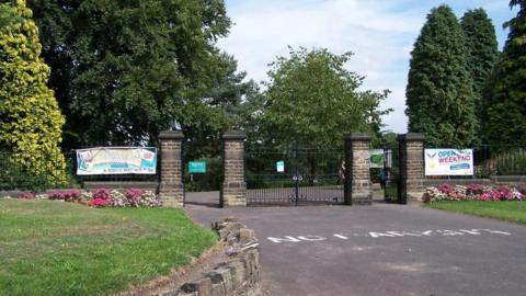 The gated entrance to Graves Park. There are four large stone pillars around 8ft tall with large cast iron gates. In front there is a tarmac road with raised verges on either side and borders of pink and white flowers.