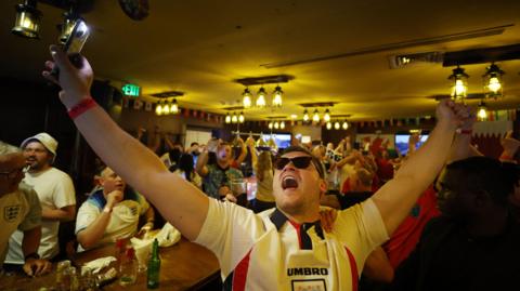 An England football fan celebrates while watching a game at a pub.
