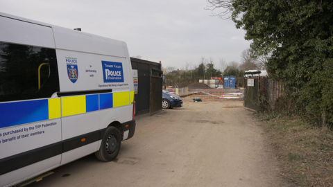 The back end of a Thames Valley Police van can be seen parked up along a wooden boarded fence, in front of the entrance to a construction site. There is a sandy dirt track, with a grass verge and some hedging and trees to the right. Through the fence, in the distance, are shipping containers, mounds of rubble and a blue car and orange cordons.