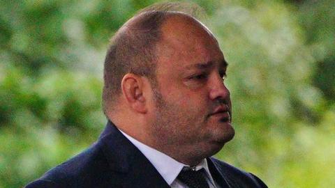 John Stringer outside court in front of greenery in the far background. He is a heavy set man, bald on top with short cropped hair on the sides and with stubble